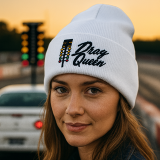 Woman wearing a white embroidered beanie featuring the drag-racing light tree and “Drag Queen” text, standing at a drag strip during sunset with cars in the background — Geaux Big motorsport lifestyle photo.