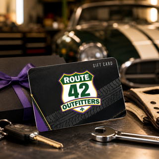 Route 42 Outfitters gift card displayed on a dark garage workbench with car keys, wrench, driving gloves, purple ribbon gift box, and a blurred classic car in the background.