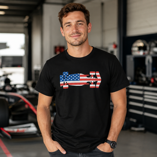 Man wearing a black unisex T-shirt featuring a USA flag Formula racing car graphic, standing in an automotive workshop with a race car in the background.